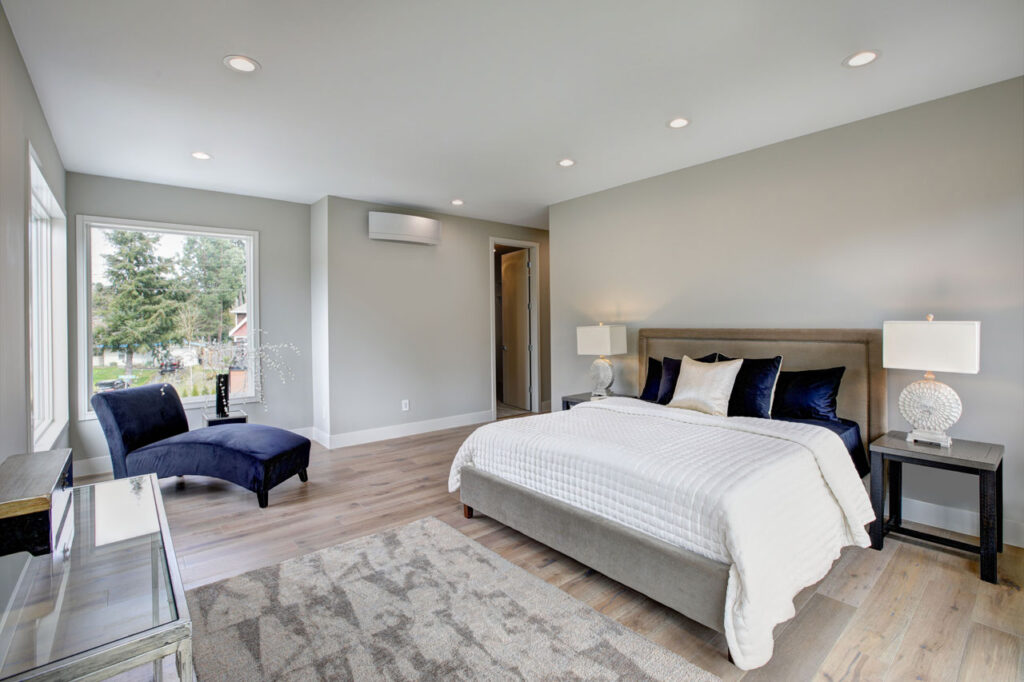 A spacious and modern bedroom with a large bed dressed in white and navy bedding, flanked by sleek nightstands with decorative lamps. The room features light wood flooring, a plush area rug, a navy velvet chaise lounge, and large windows offering a view of the outdoors.