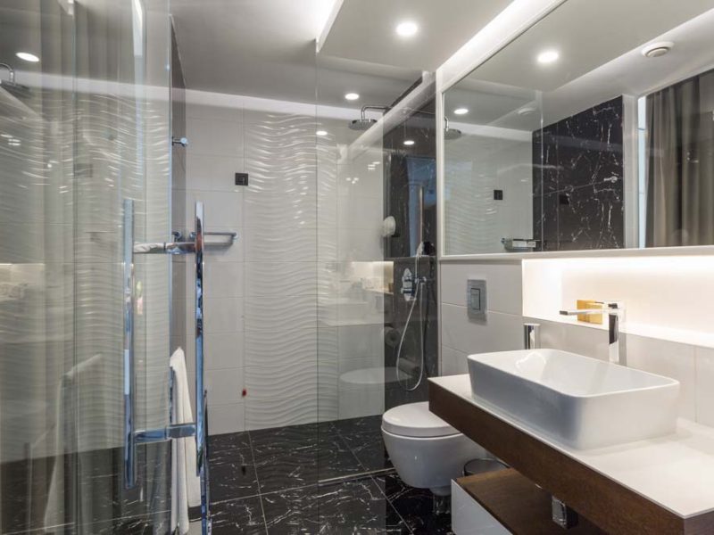 A sleek contemporary bathroom with a glass-enclosed shower, wave-patterned wall tiles, and a dark marble floor. Features include a vessel sink on a wooden vanity, a wall-mounted toilet, recessed lighting, and a large mirror with ambient backlighting for a luxurious feel.