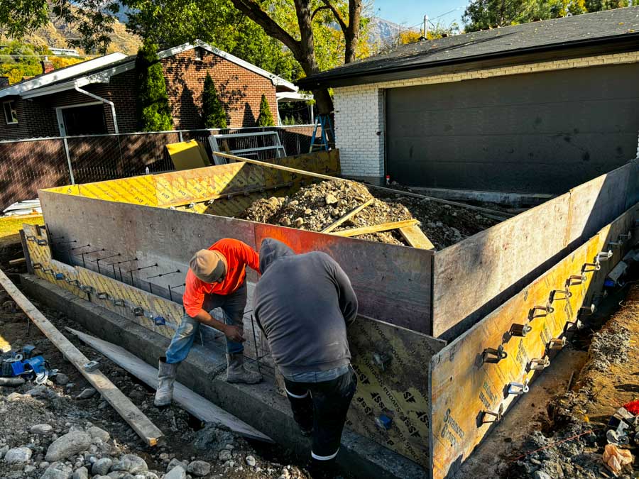 Two construction workers are bent over, securing rebar within wooden foundation forms filled with gravel. The site is surrounded by building materials and tools, with a garage and neighboring homes visible in the background, indicating early-stage home construction.