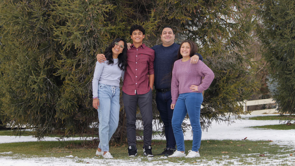 A full-length color photo of a family of four standing together outdoors in a snowy park. They are positioned in front of evergreen trees, with patches of snow on the ground. The group is casually dressed, smiling, and appears relaxed and happy.